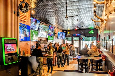 Busy sports bar with patrons at the counter, multiple TVs, neon signs, and warm industrial decor