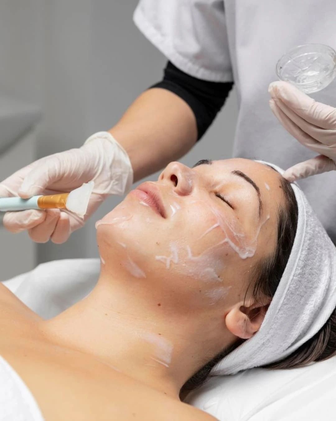 Persona recibiendo tratamiento facial; cosmetóloga aplicando una crema con una brocha. Ajuste blanco.