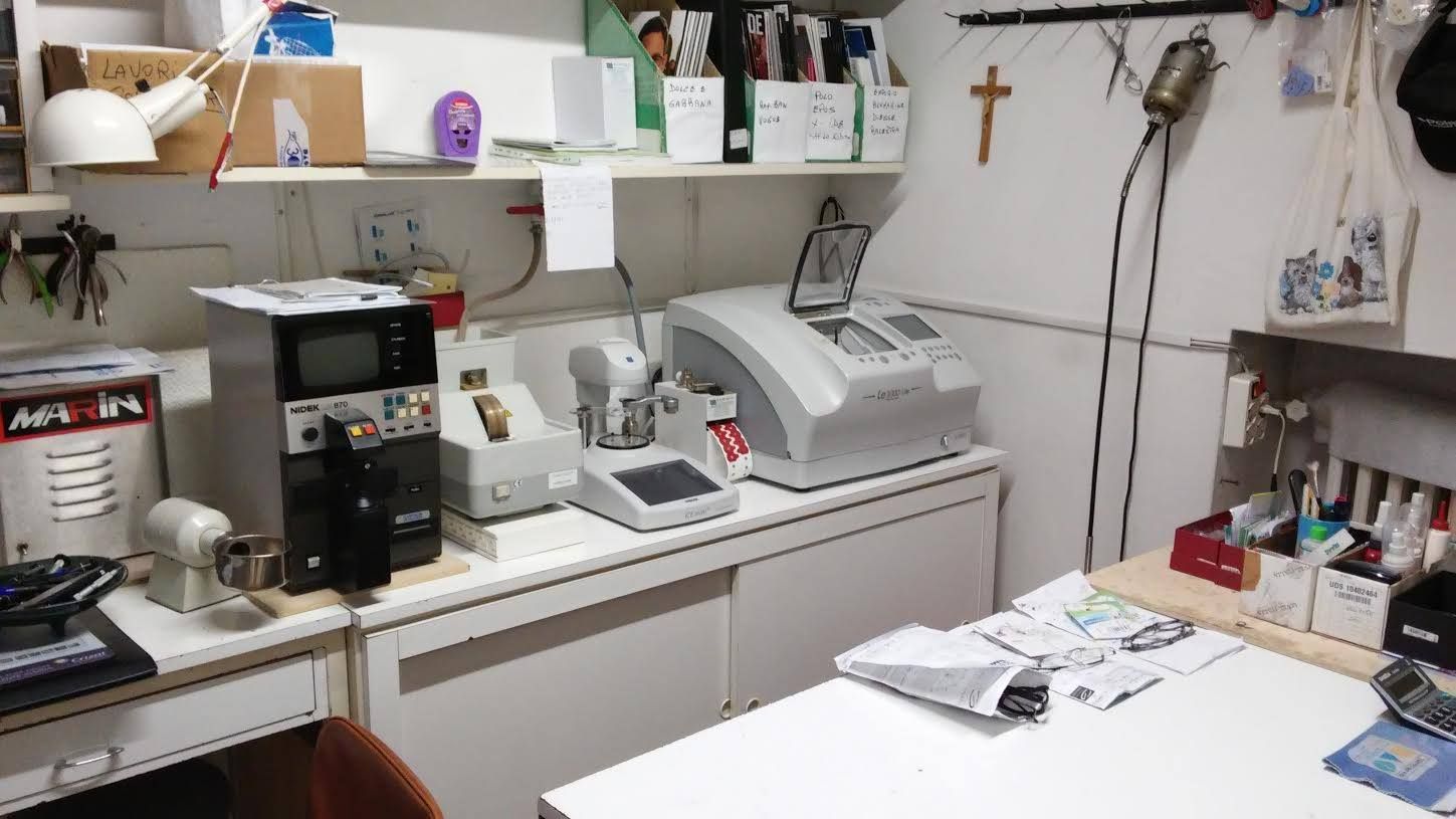 Un paio di occhiali geometrici esposti su uno scaffale rosa in un negozio. Montatura nera con lenti trasparenti e un'immagine promozionale sullo sfondo.