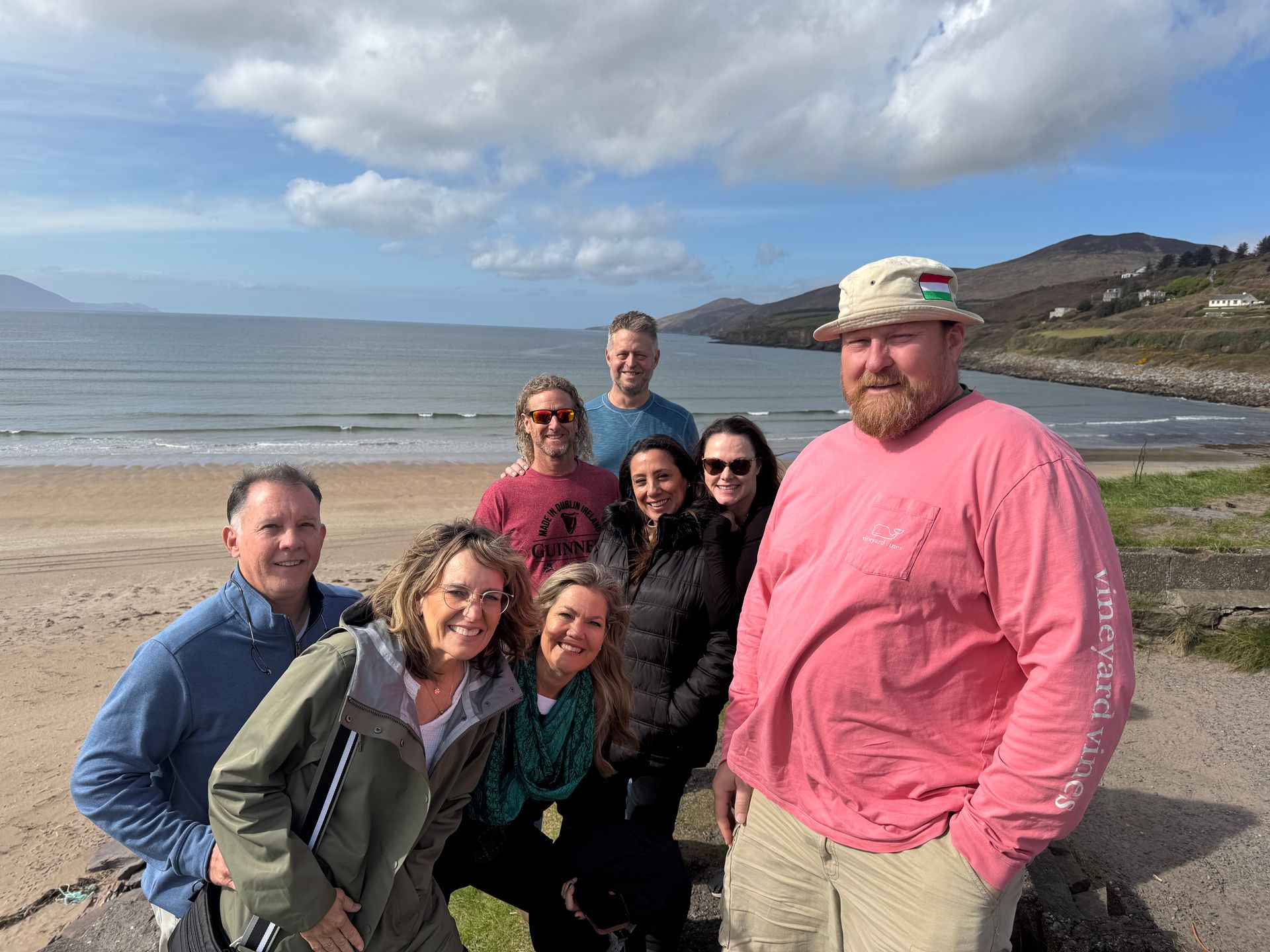 Group of six smiling people posing on a coastal road with ocean, beach, and hills in the background near dingle