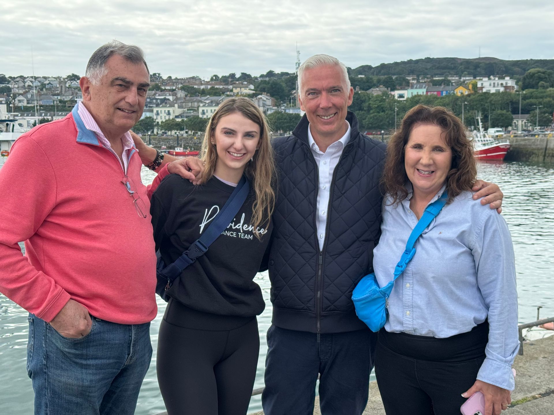 Four people pose by water, arms around each other. Background has buildings, boats, and hills under an overcast sky.