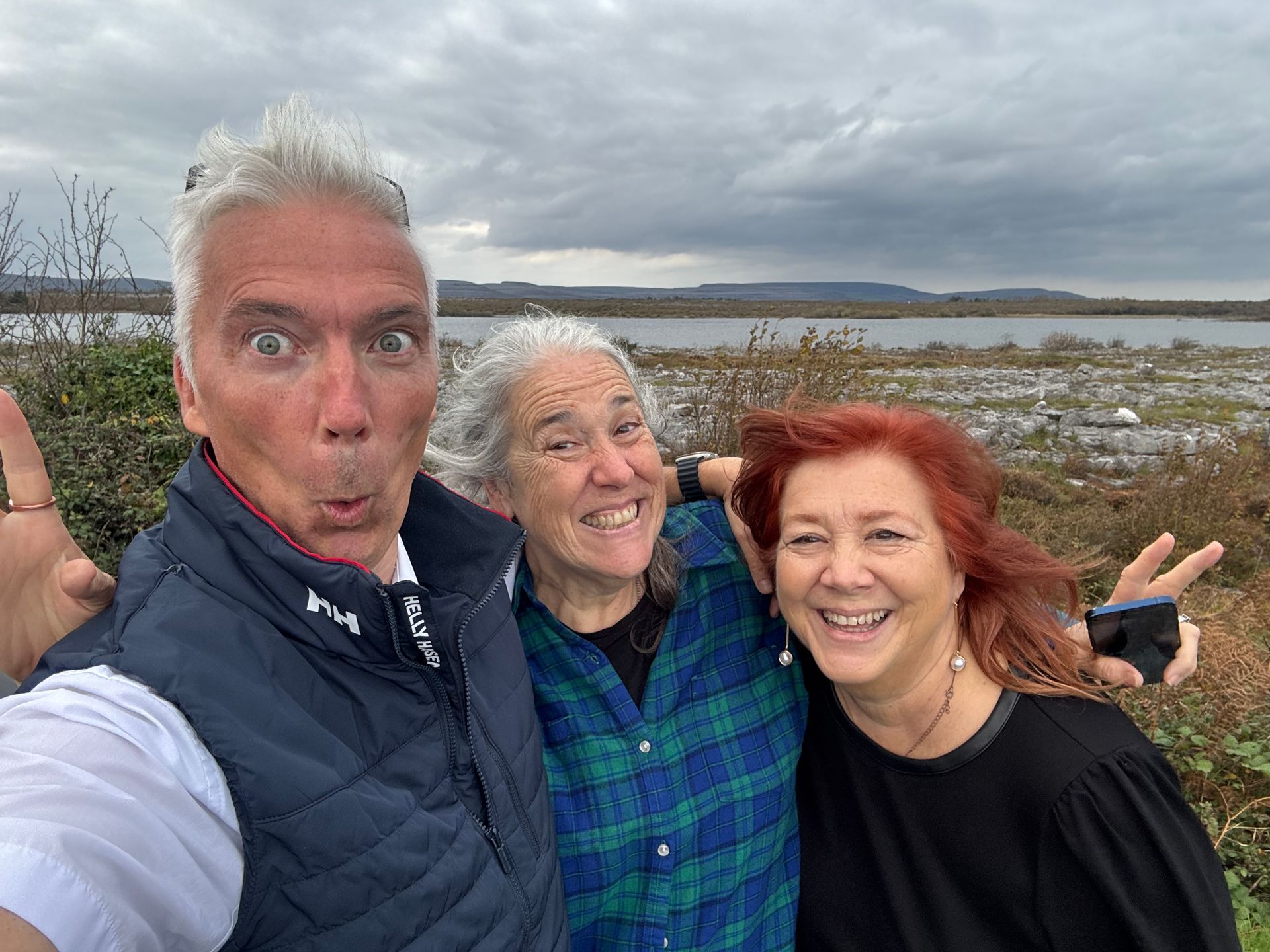 Three people pose outdoors near water, making silly faces. Cloudy sky, two women, one man.