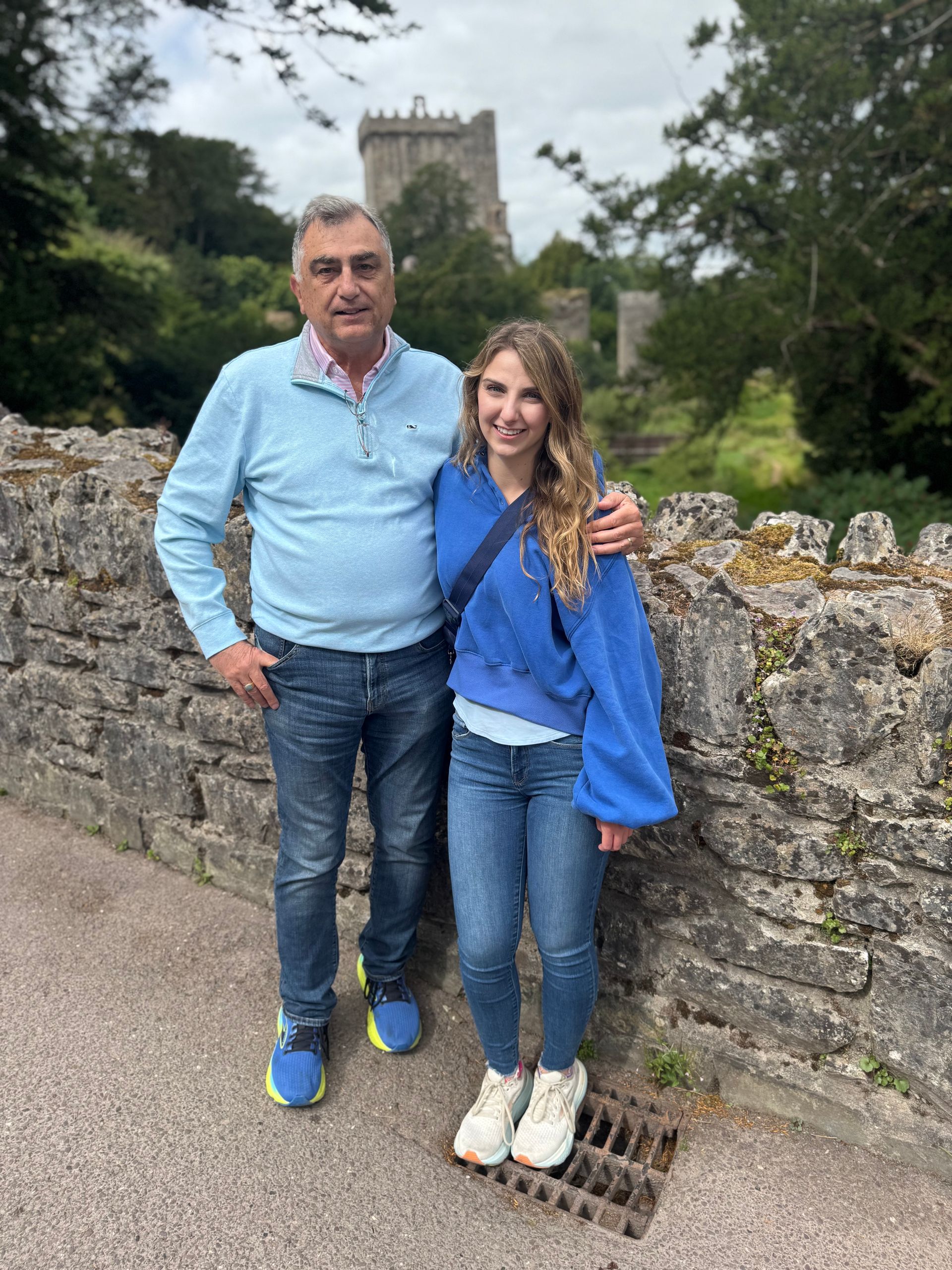 Man and woman pose on stone bridge with castle in background. Man has arm around woman.