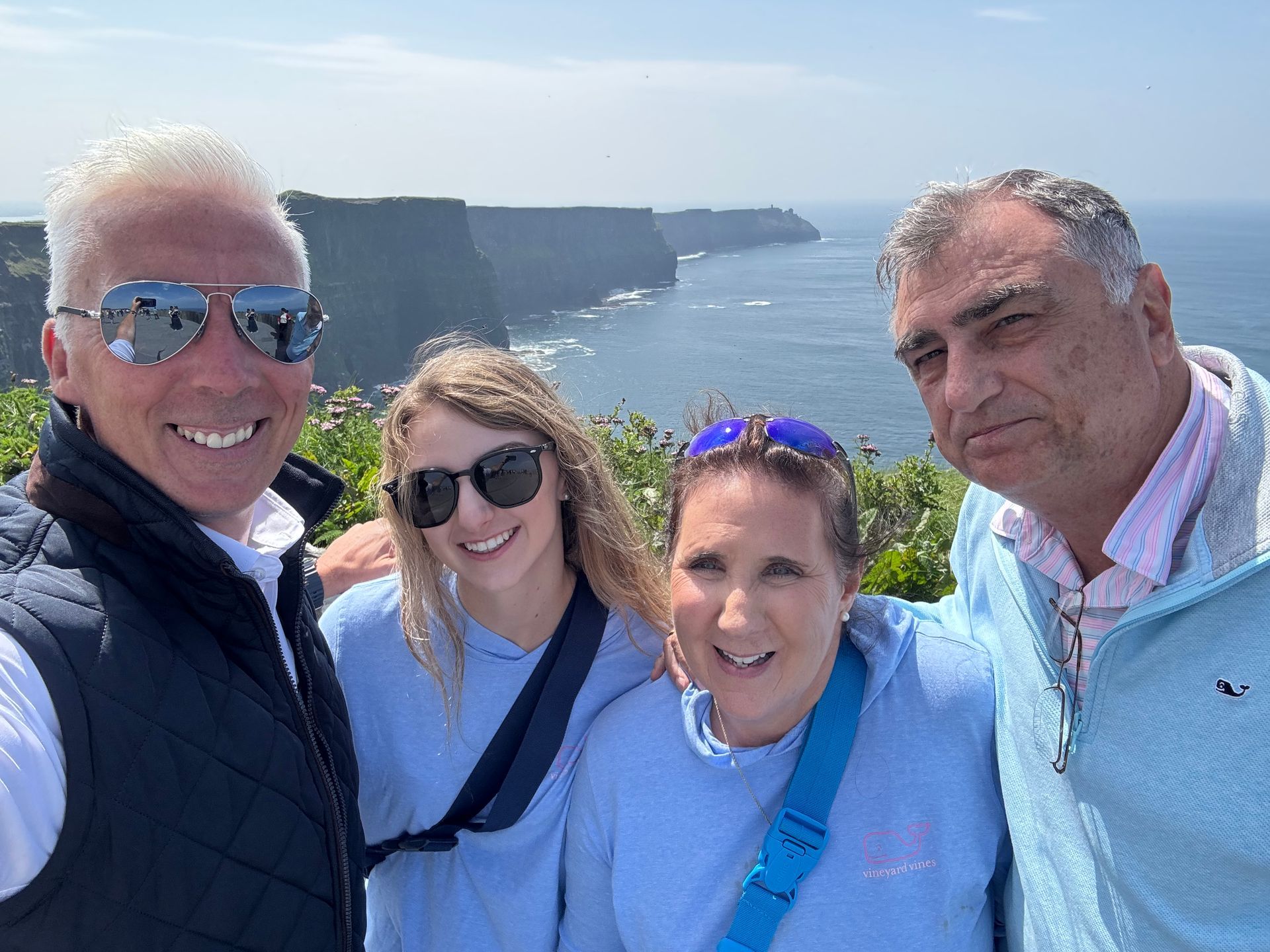 Four smiling friends take a selfie by a sunny seaside cliff.