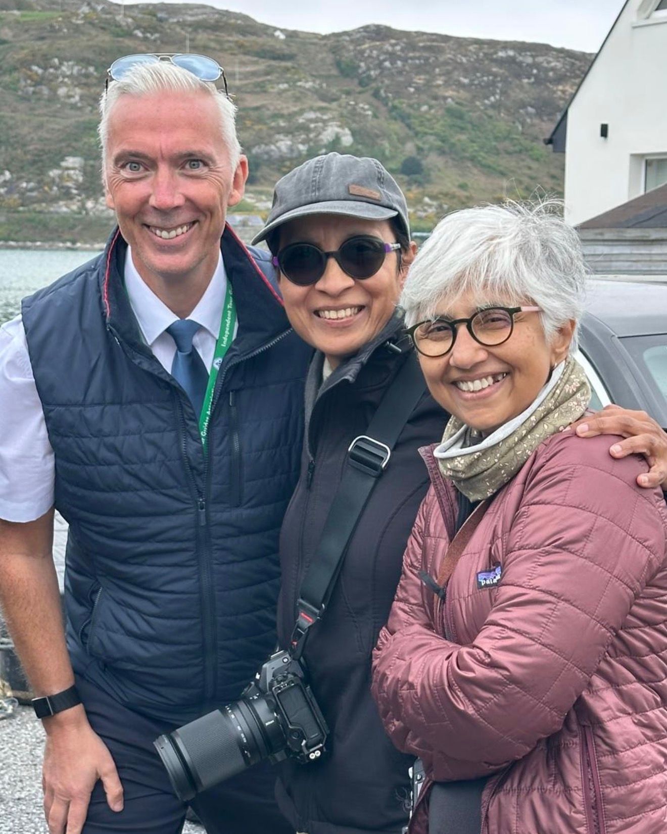 Three smiling people outdoors with a camera, standing beside a car in a hilly area.