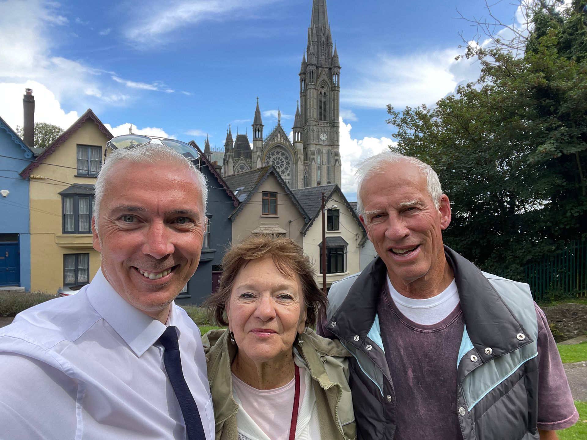 Three people smiling in front of historic buildings and a church spire in Cobh under a blue sky