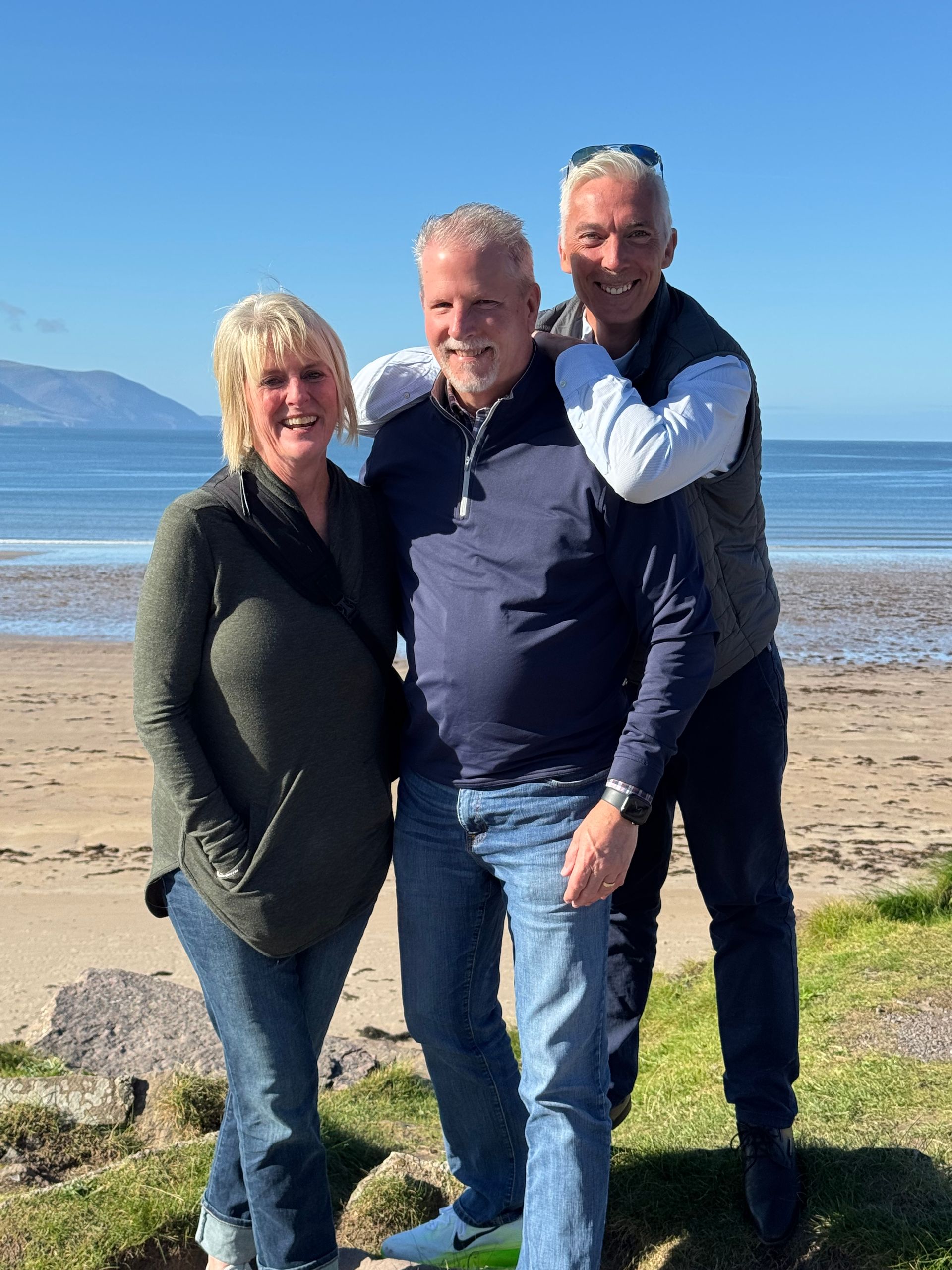 Three people smiling on a beach; sunny day. Woman in green sweater, men in blue shirts.