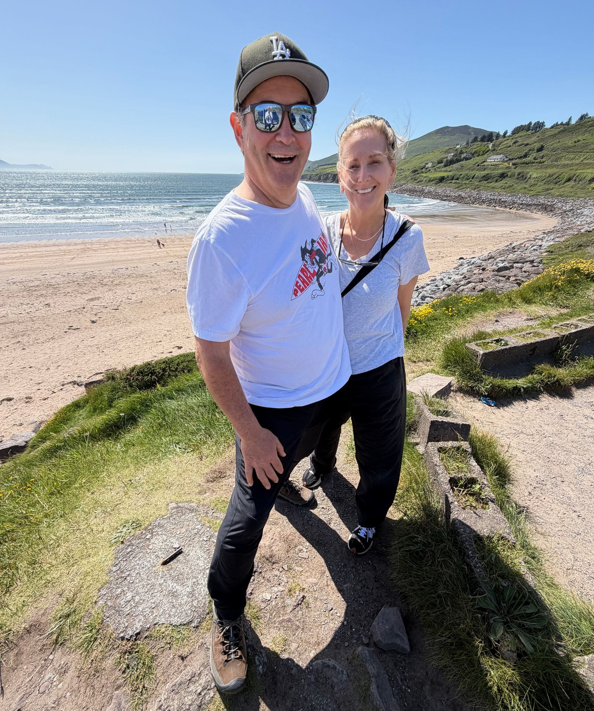 Two people smiling on a sunny beach path beside the ocean, with waves and hills in the background.