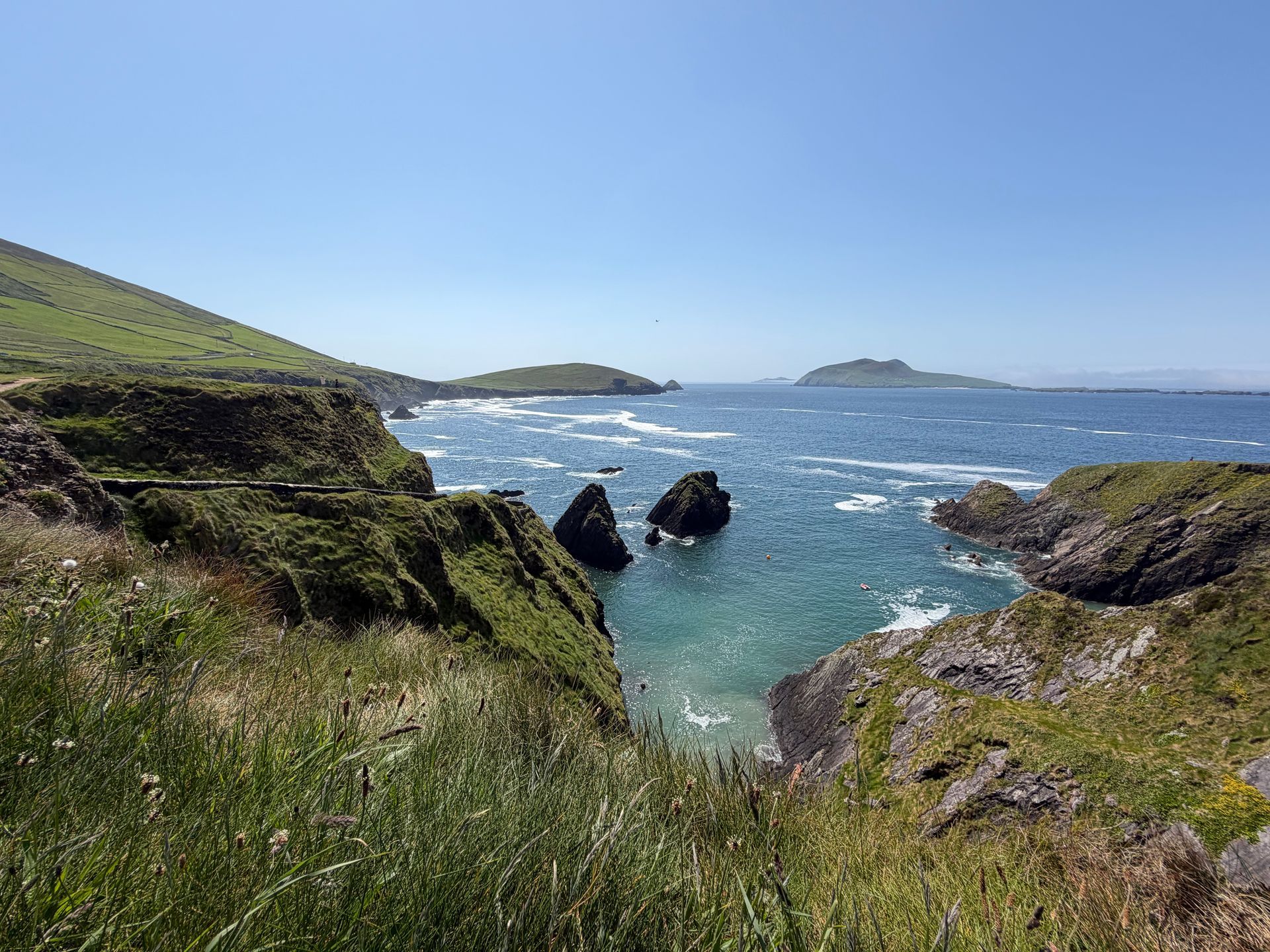 Rocky coastal cove with turquoise water, grassy cliffs, and distant islands under a clear blue sky