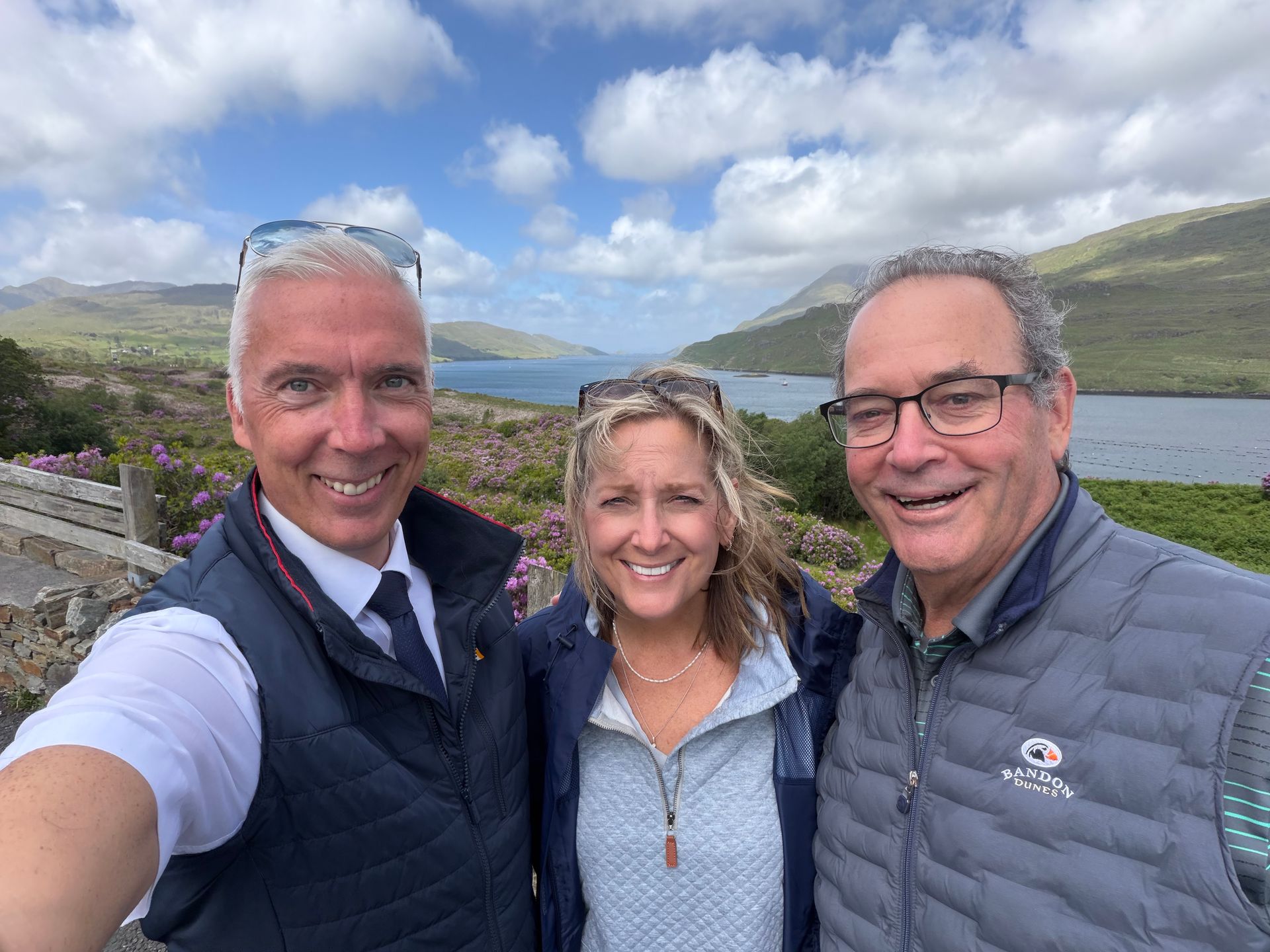 Three people smiling for a selfie by a lake with hills and cloudy sky in the background