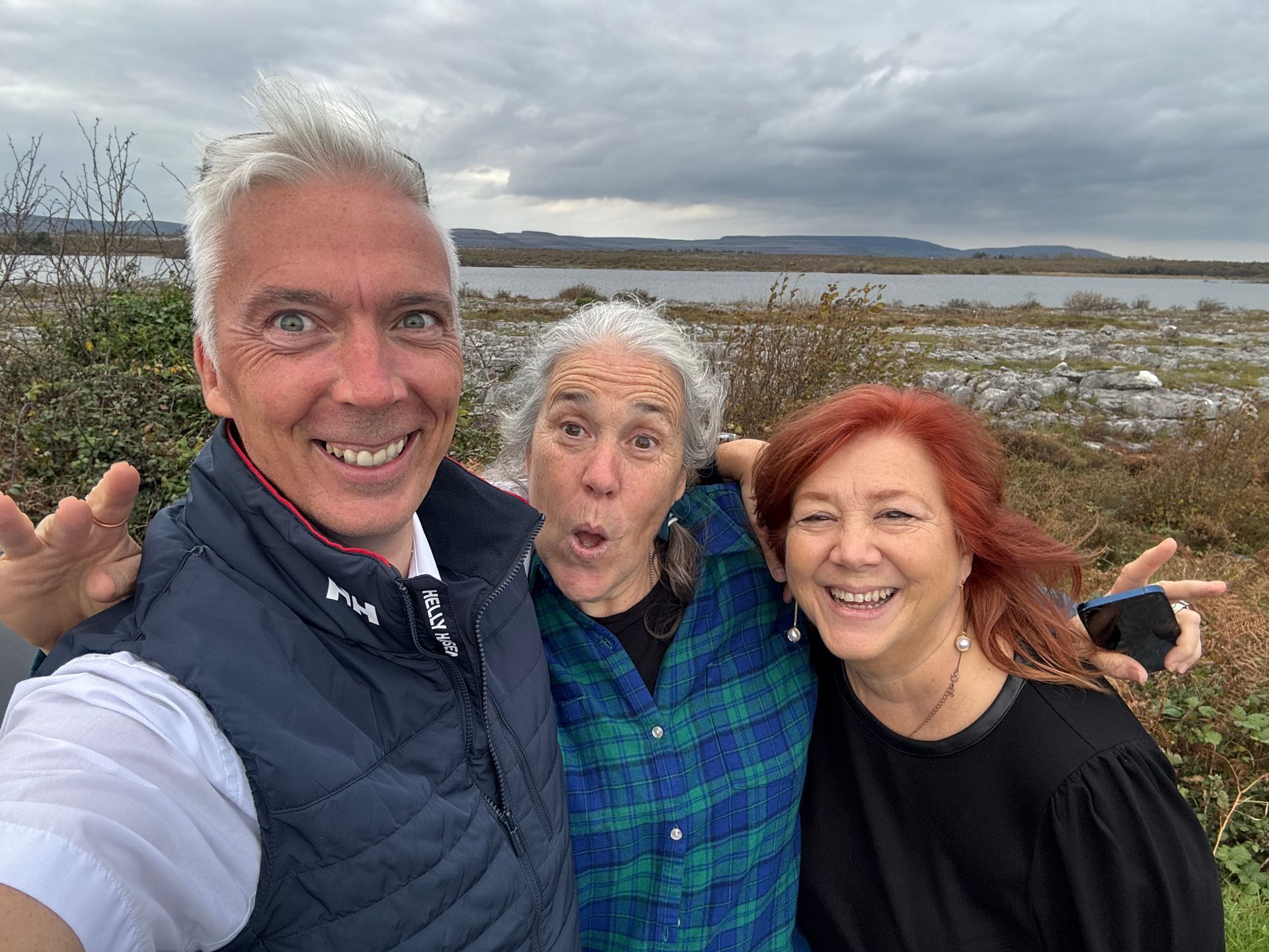 Three smiling people posing for a selfie outdoors near water under a cloudy sky