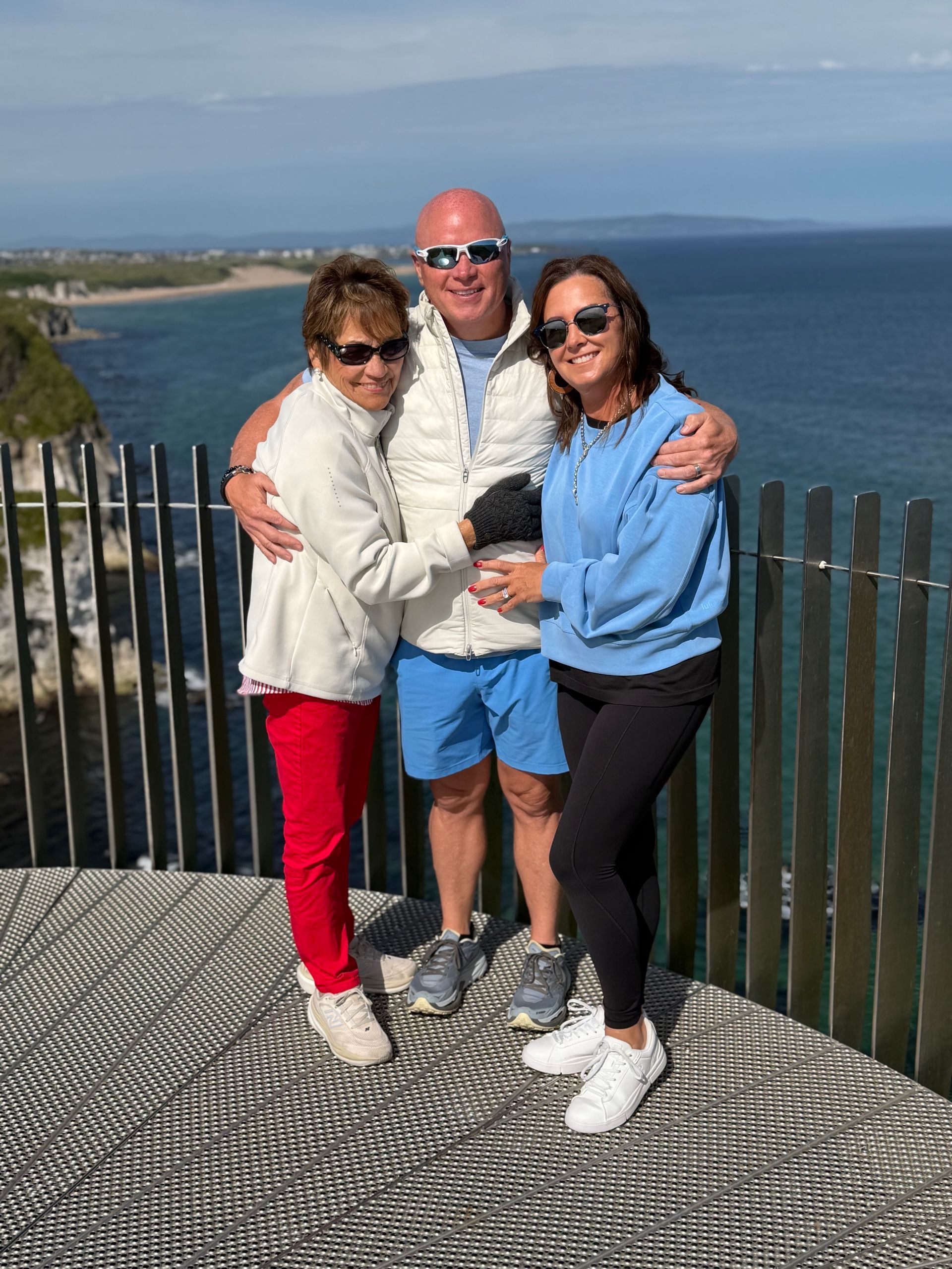 Three people posing together by a seaside overlook, smiling with ocean and cliffs in the background.