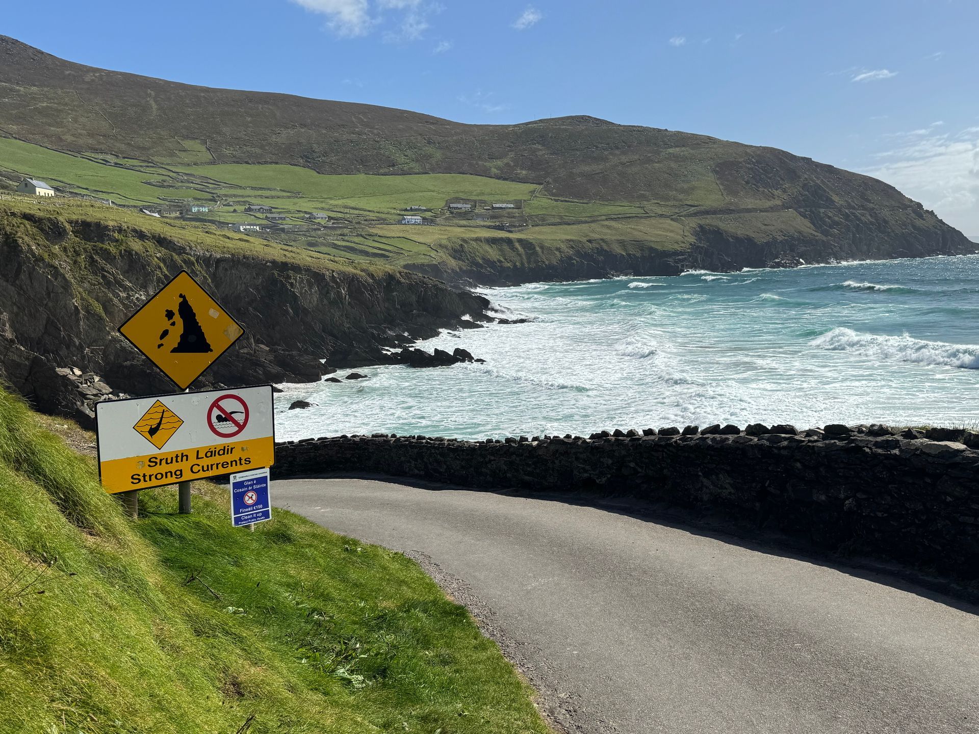 Road alongside a rocky coastline, warning signs in foreground. Green hillside and turbulent ocean under a blue sky.
