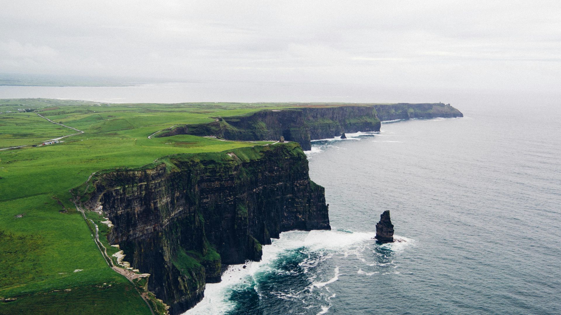 Green coastal cliffs of Moher beside a calm gray sea, with a rocky sea stack offshore.