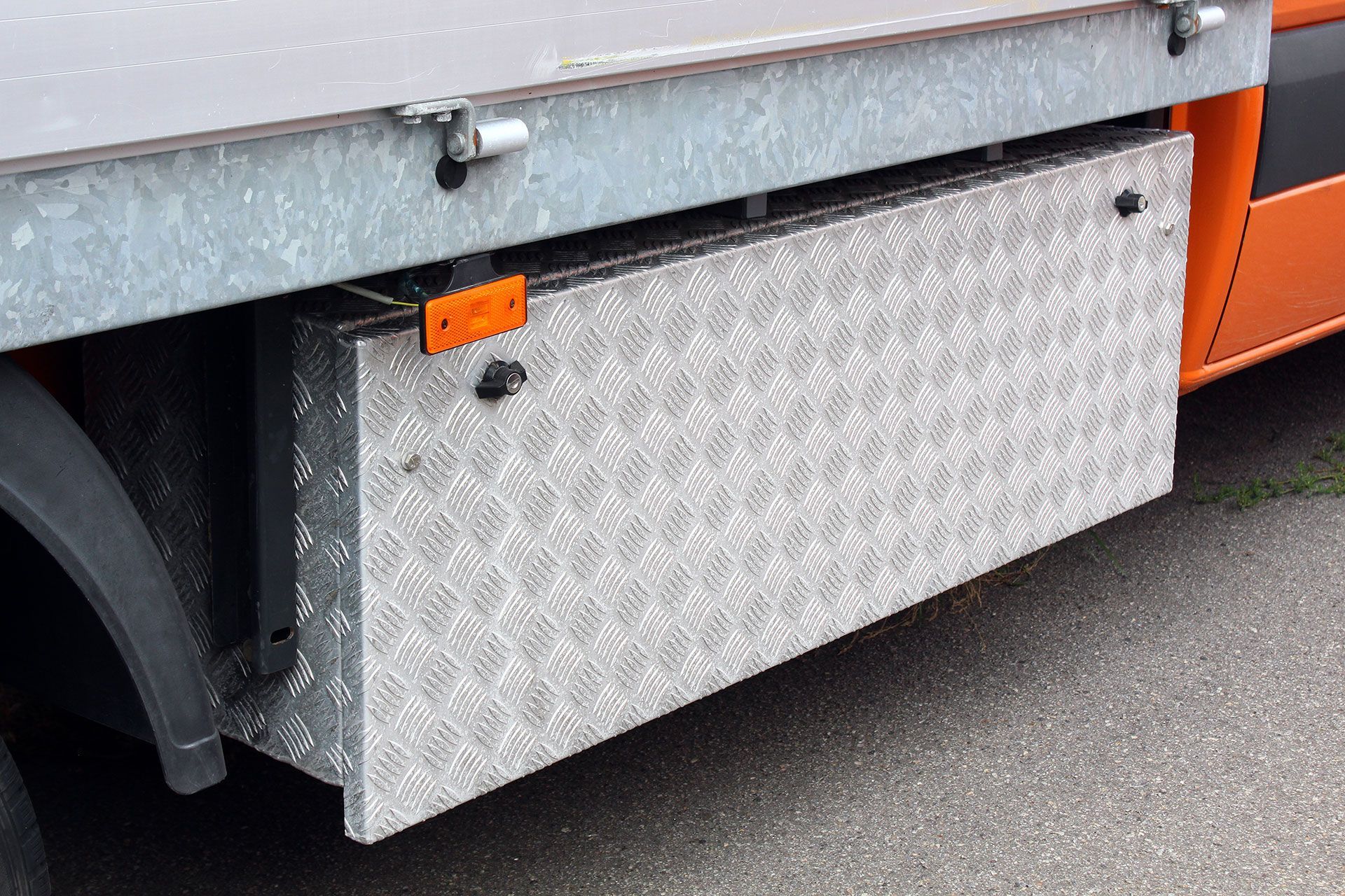 A close-up of truck metal toolbox with a ribbed plate mounted underneath a truck bed. The van tool box has a silver diamond pattern, black locks, and an orange reflector.