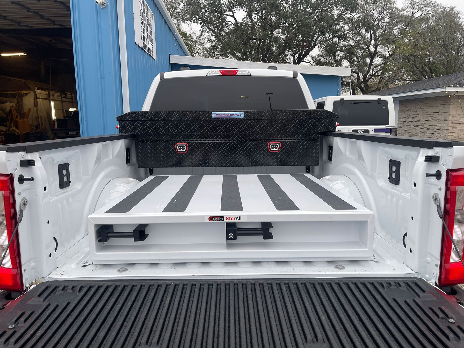 A close-up of truck metal toolbox with a ribbed plate mounted underneath a truck bed. The van tool box has a silver diamond pattern, black locks, and an orange reflector.