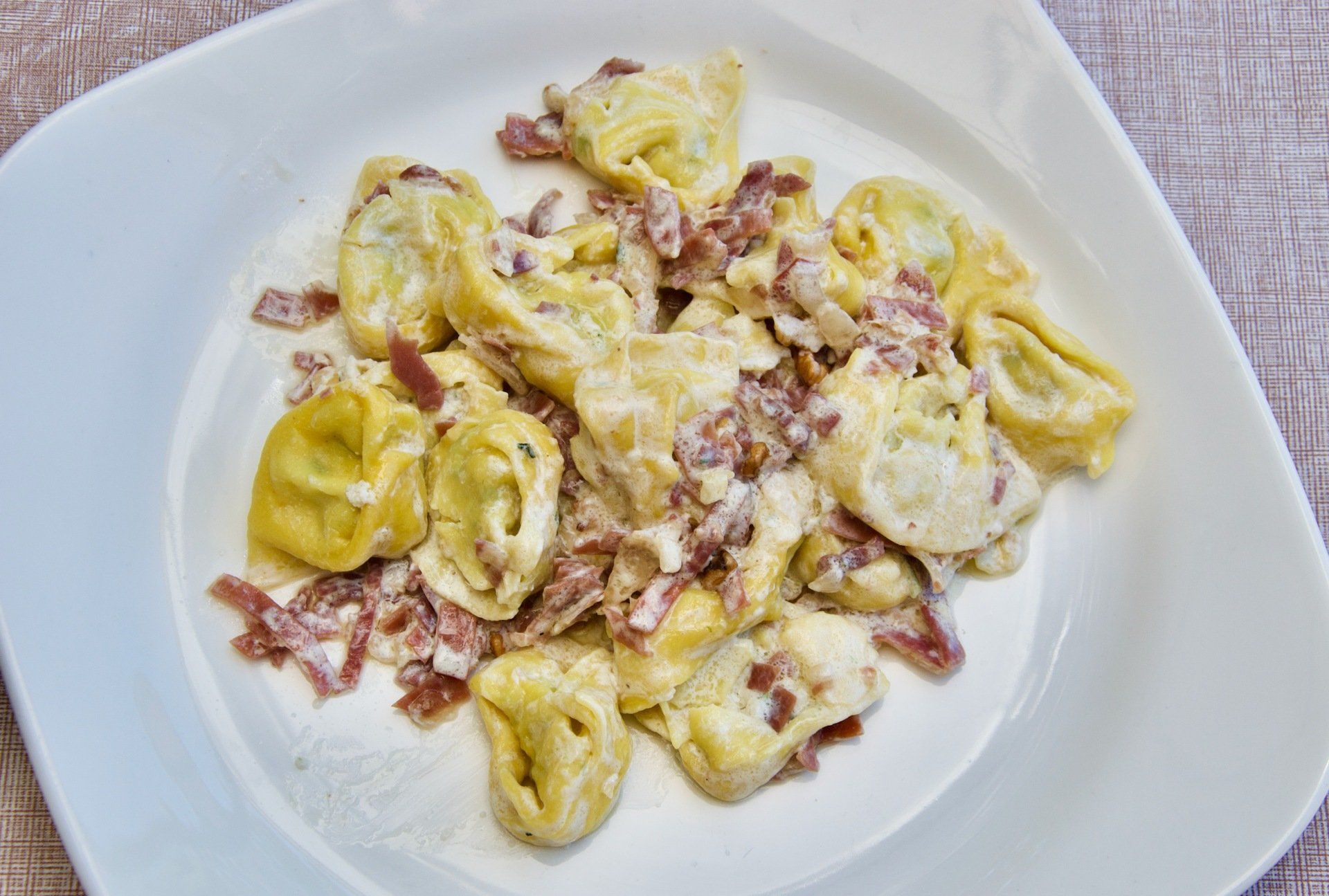 Un piatto bianco con tortellini e pancetta su un tavolo.