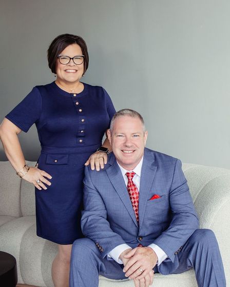 Woman in blue dress and man in blue suit pose on a couch; she rests a hand on his shoulder.