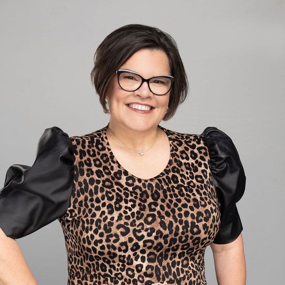 Woman with dark hair and glasses smiles, wearing leopard-print top with black puffy sleeves.