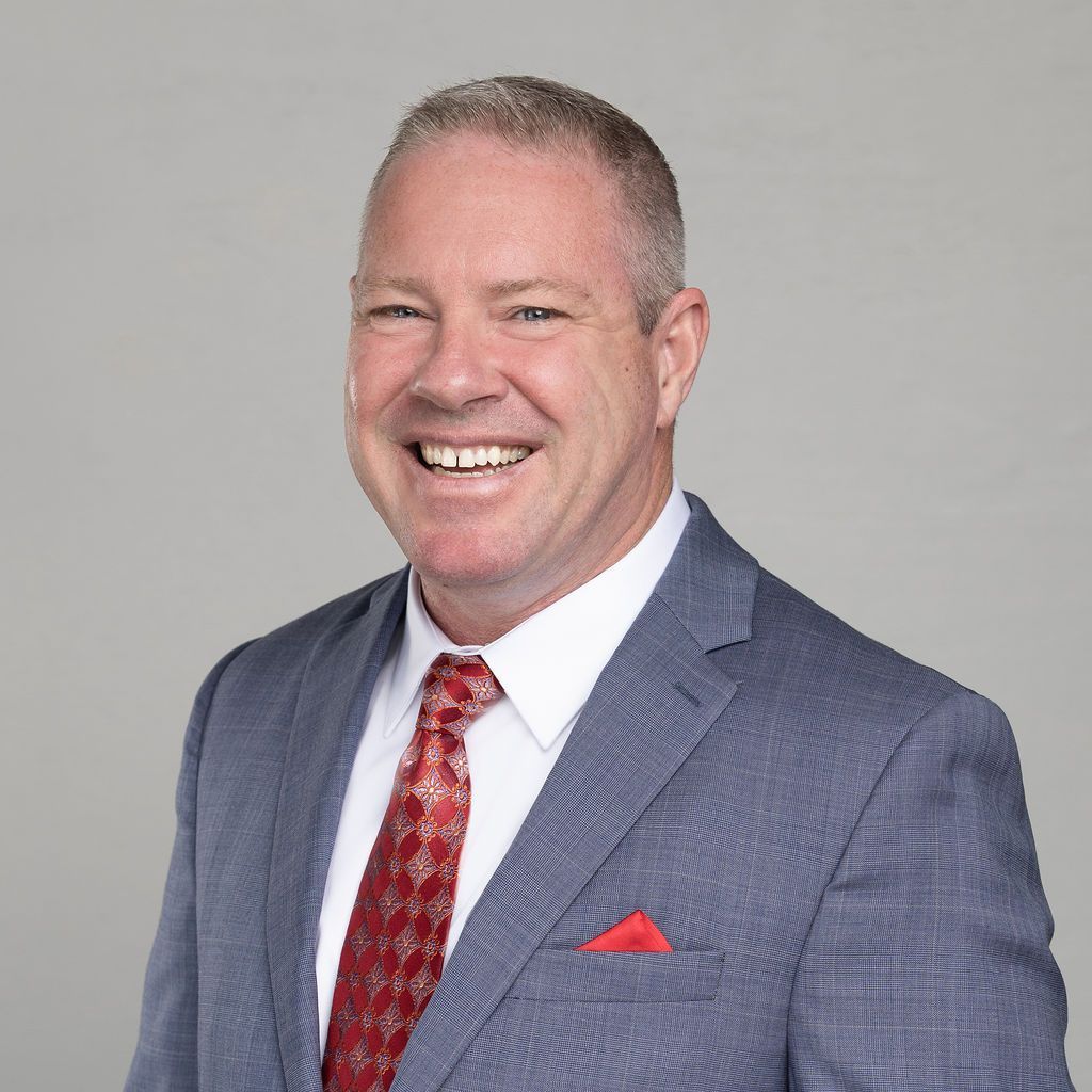 Smiling man in a blue suit, red tie and pocket square against a gray backdrop.