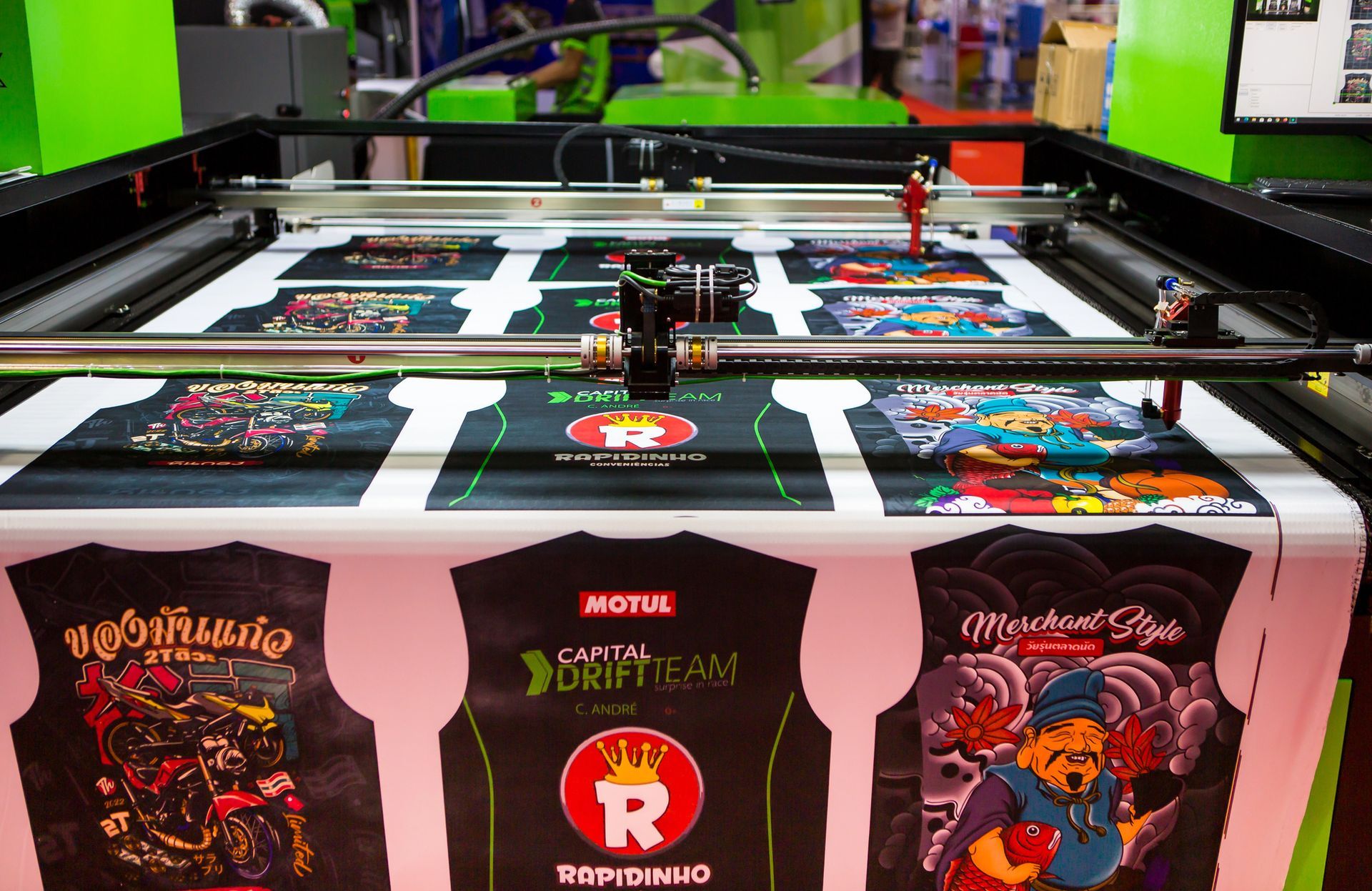 Una impresora digital cortando diseños de camisetas en un taller. Máquina negra y verde con impresiones gráficas a color.