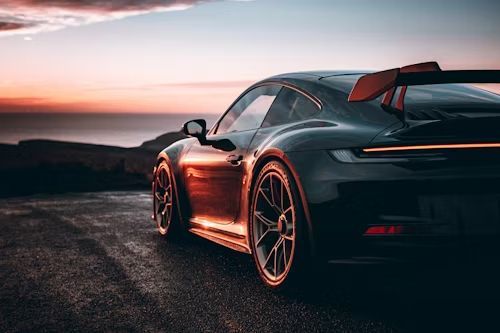 Black Porsche sports car parked on a road overlooking the ocean at sunset.