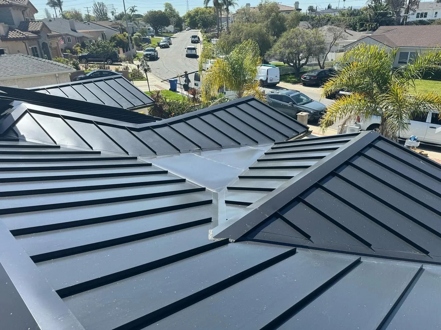 Dark gray metal roof with ridgelines and valley. Houses and street in the background.