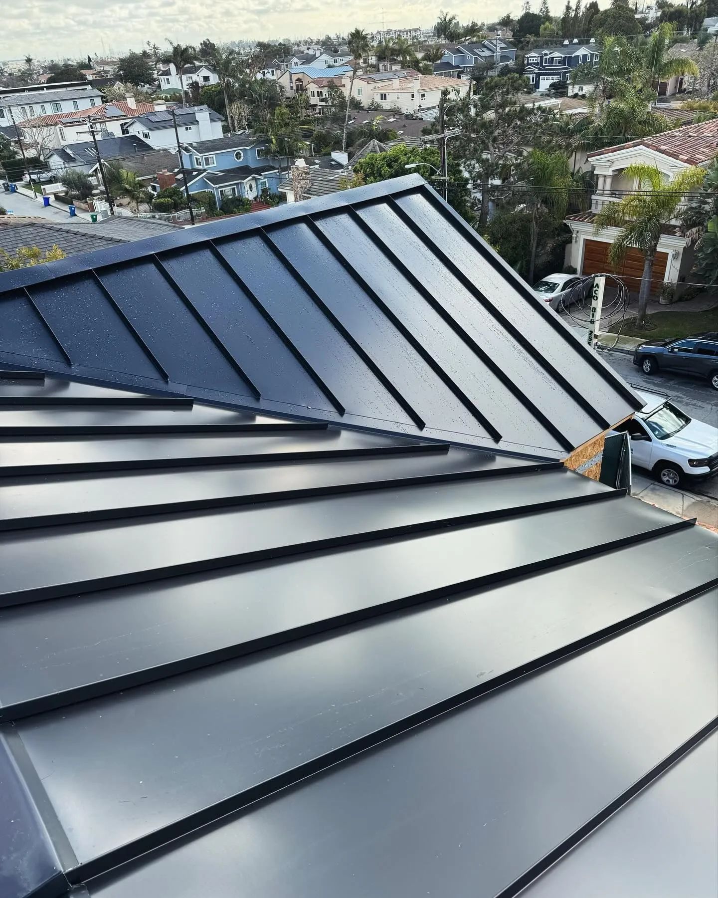 A high-angle view of a newly installed dark metal standing seam roof on a sunny day overlooking a residential neighborhood.