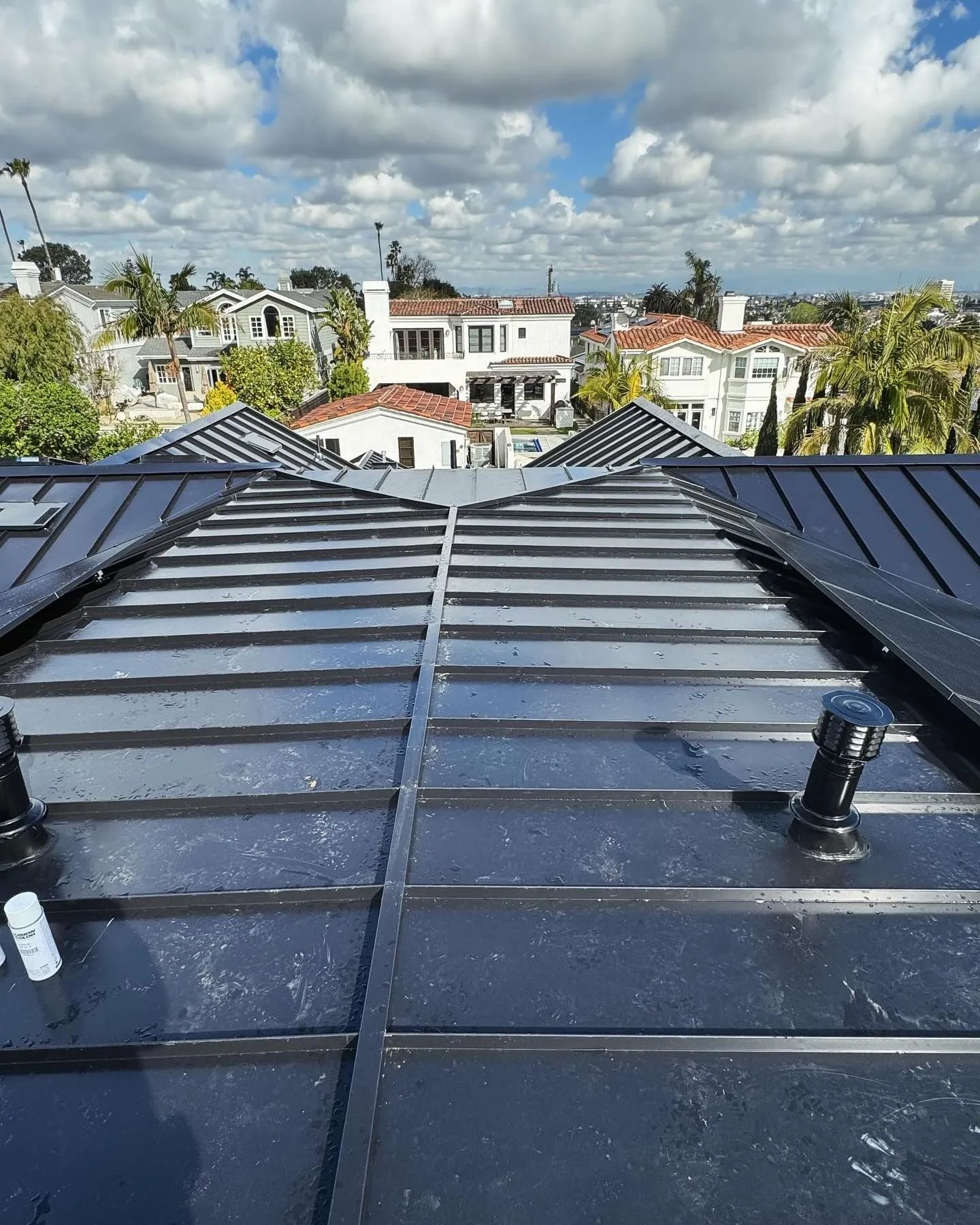 Black metal roof with solar panels, a sunny sky, and houses in the background.