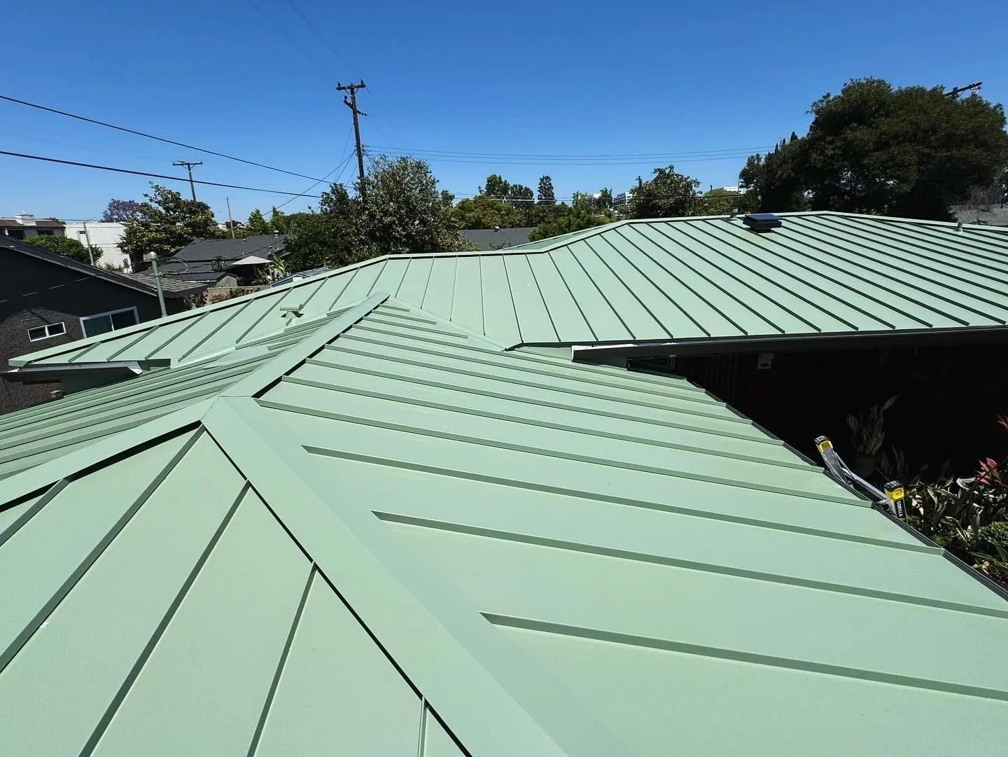 Green metal roof on a house under a bright blue sky, with power lines and trees in the background.