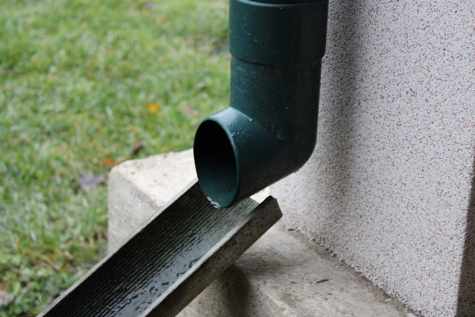 Black downspout draining water onto a sloped splash block beside a house foundation