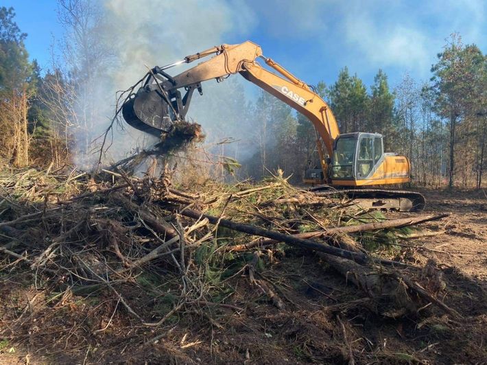 Orange excavator clearing brush in a forest, smoke rising.