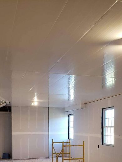 Interior of a room under construction with drywall and unfinished ceiling. Two windows and a scaffold are visible.