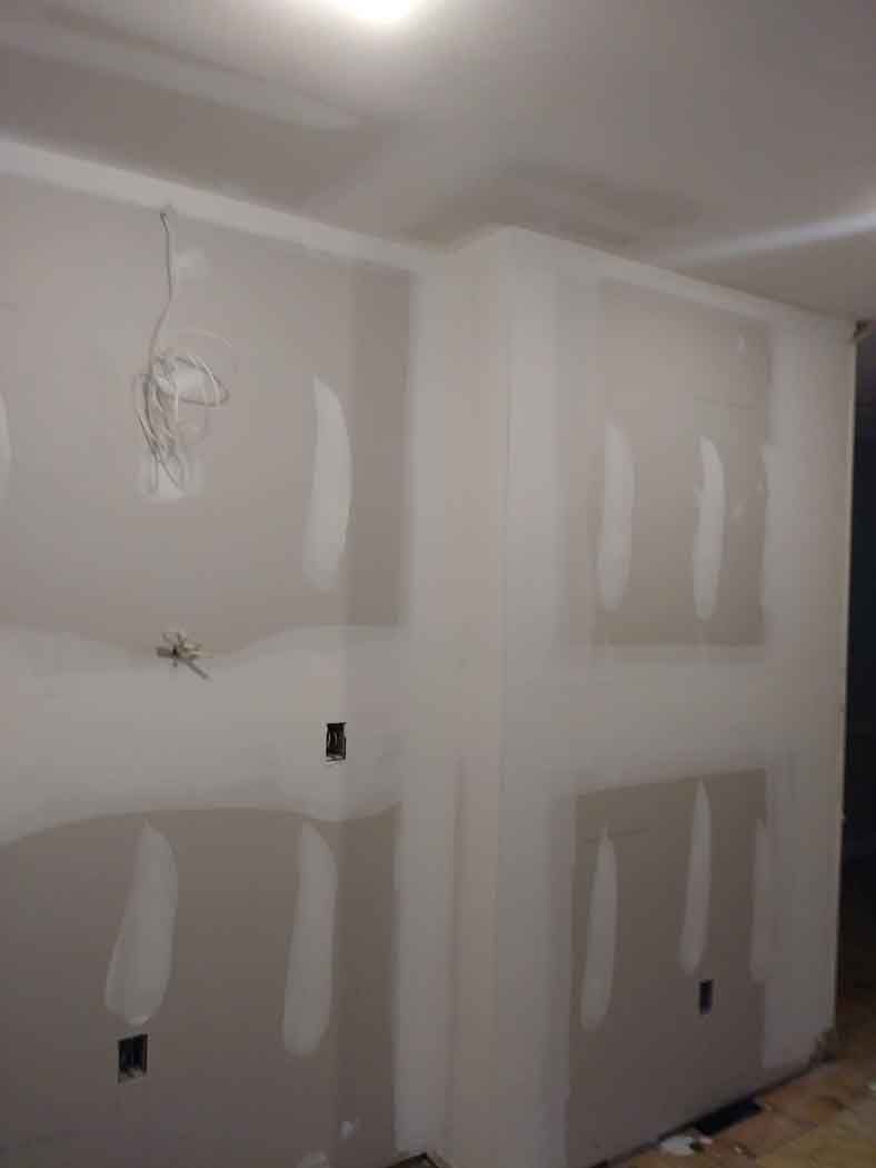 Interior corner of a room, drywall installed with taped seams and electrical boxes visible.