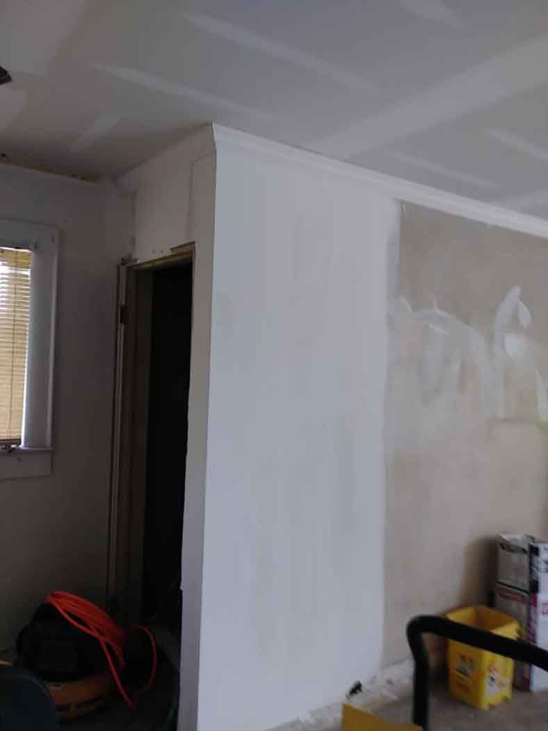 Room under renovation with drywall and unfinished walls. Doorway, electrical cords, and a yellow bucket are visible.