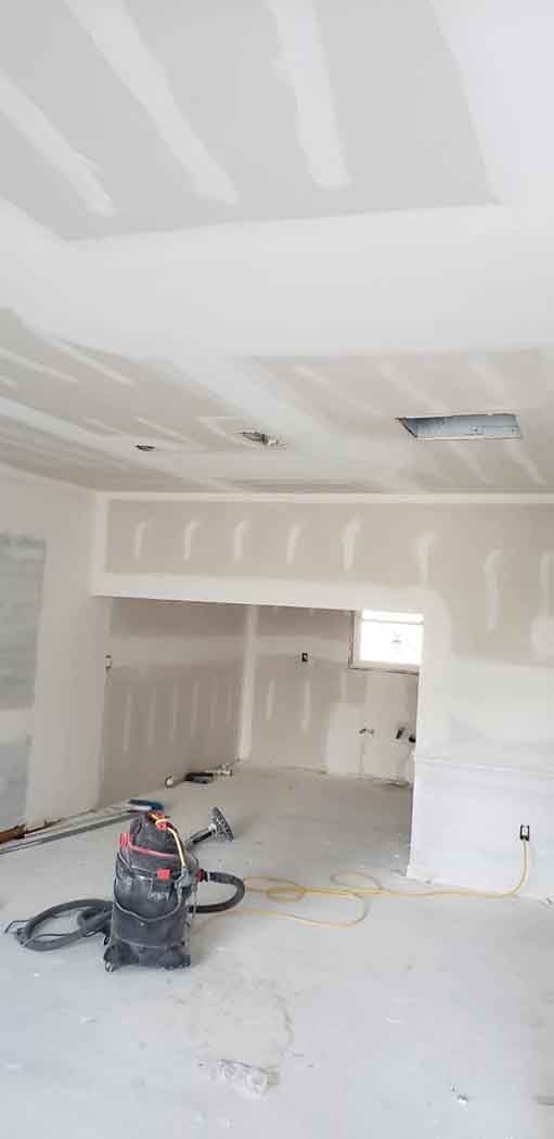 Interior of a room under construction with drywall installed on walls and ceiling. A vacuum cleaner sits on the floor.