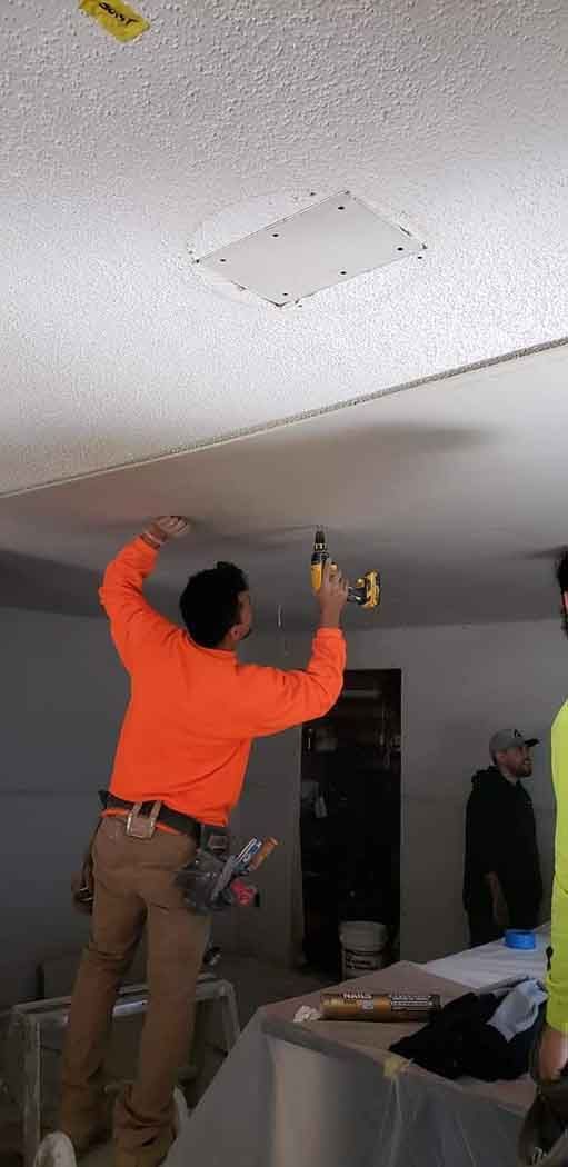 Construction worker in orange shirt drilling a ceiling panel. Other workers visible in the background.