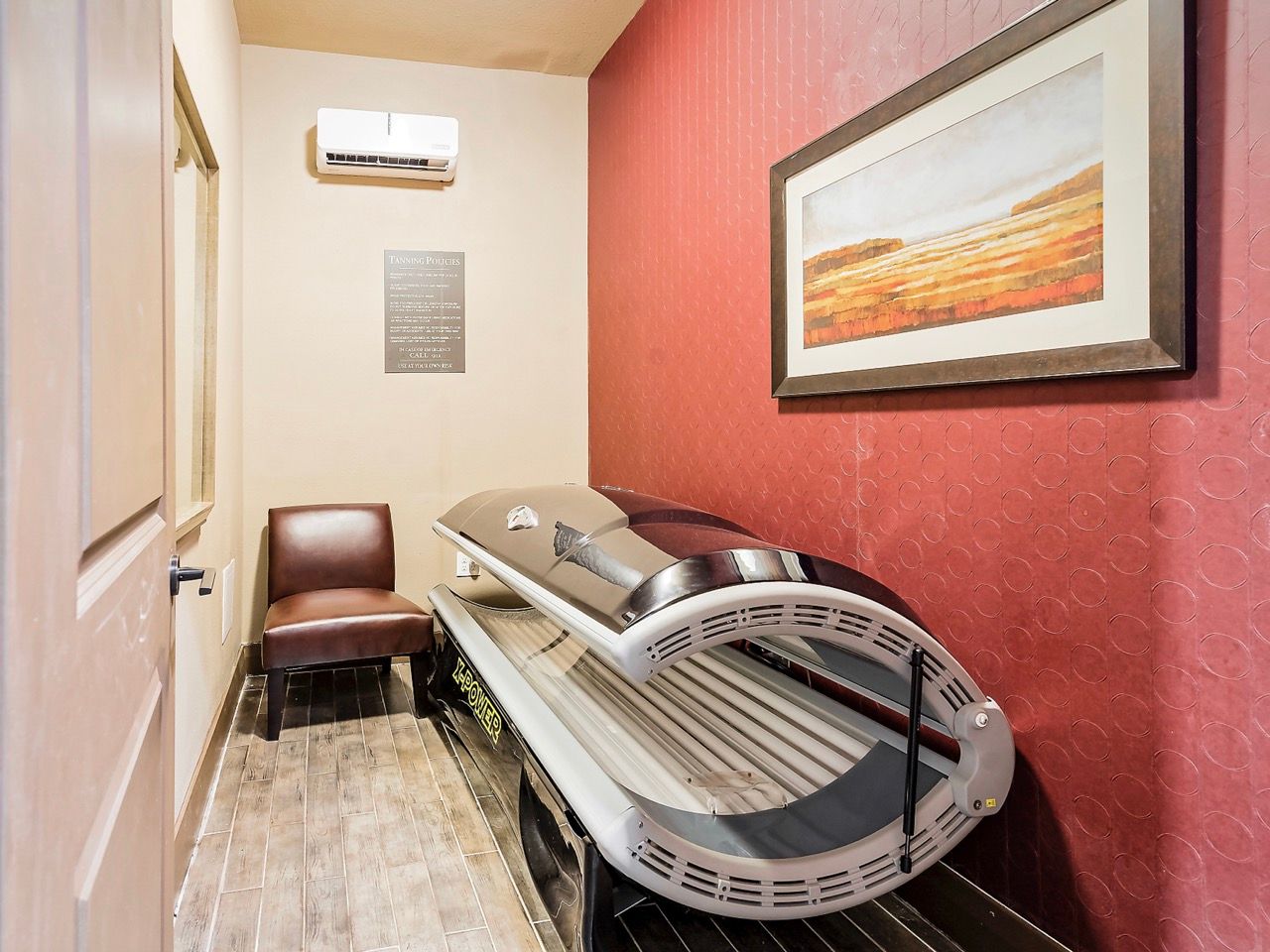 Tanning bed in a compact private room with a brown chair and red patterned wall.