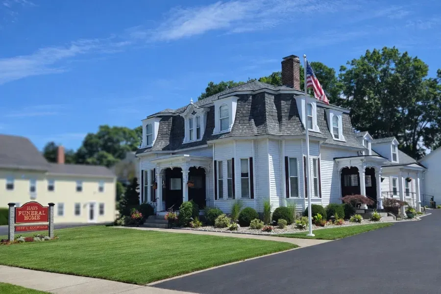 A white, multi-story historic house with a mansard roof and an American flag, set behind a lawn and a red roadside sign.