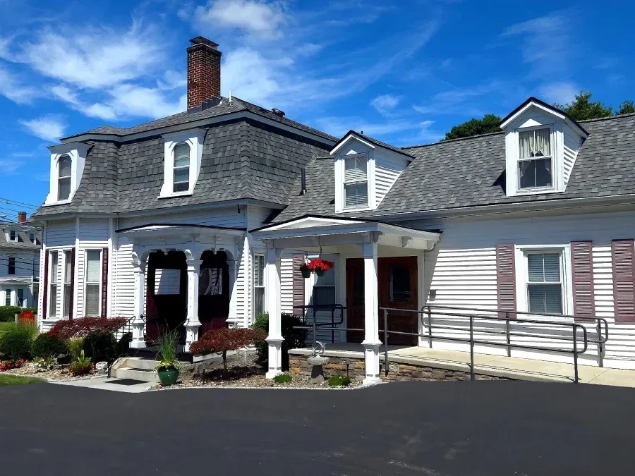 A white, two-story house with a grey shingled roof, a chimney, dormer windows, a front porch, and a wheelchair ramp.