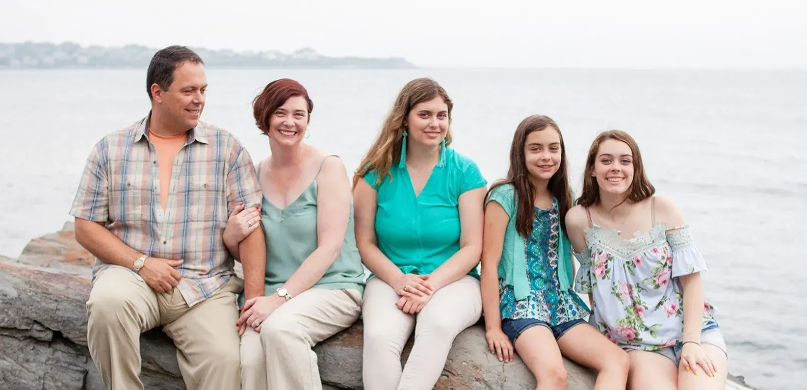 A family of five sitting together on coastal rocks, smiling toward the camera with the ocean in the background.