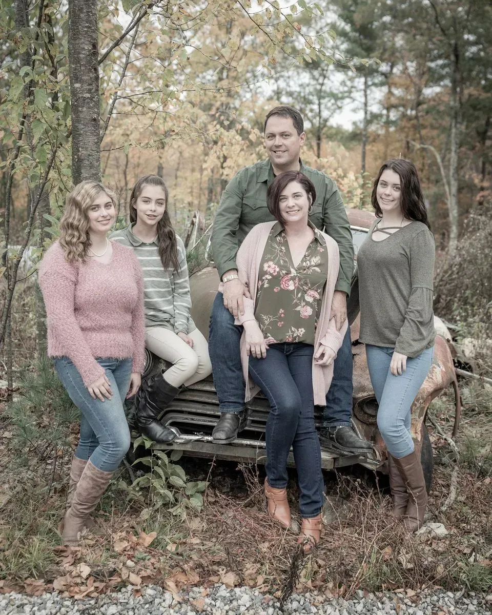 A family of five poses for a portrait in front of a rusted vintage truck in a wooded area during autumn.