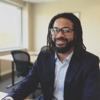 Man with locs and glasses smiles, wearing a navy blazer and light blue shirt, in an office.