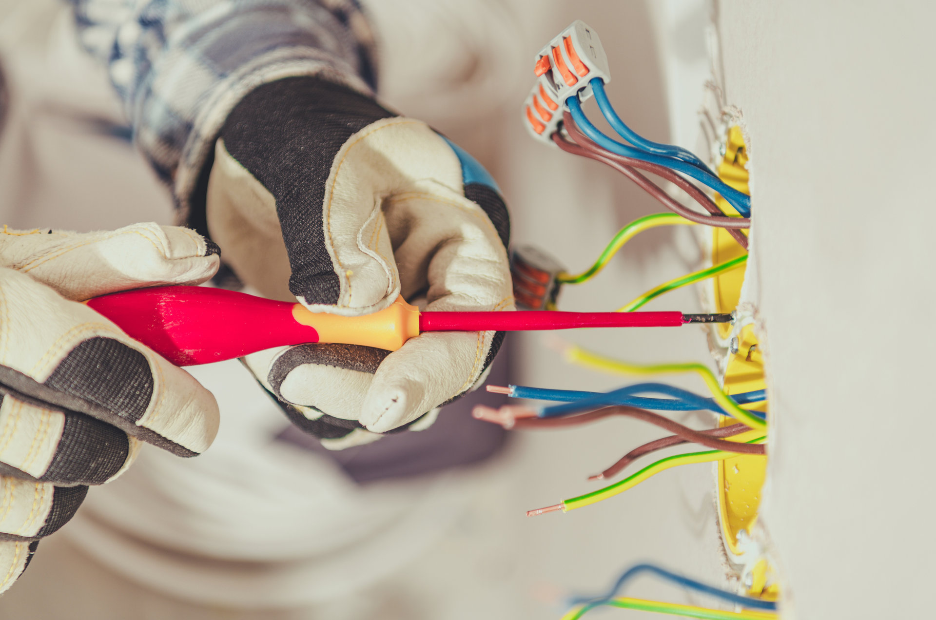 Electrician in blue coveralls and yellow hard hat inspecting electrical panel, using laptop outdoors.