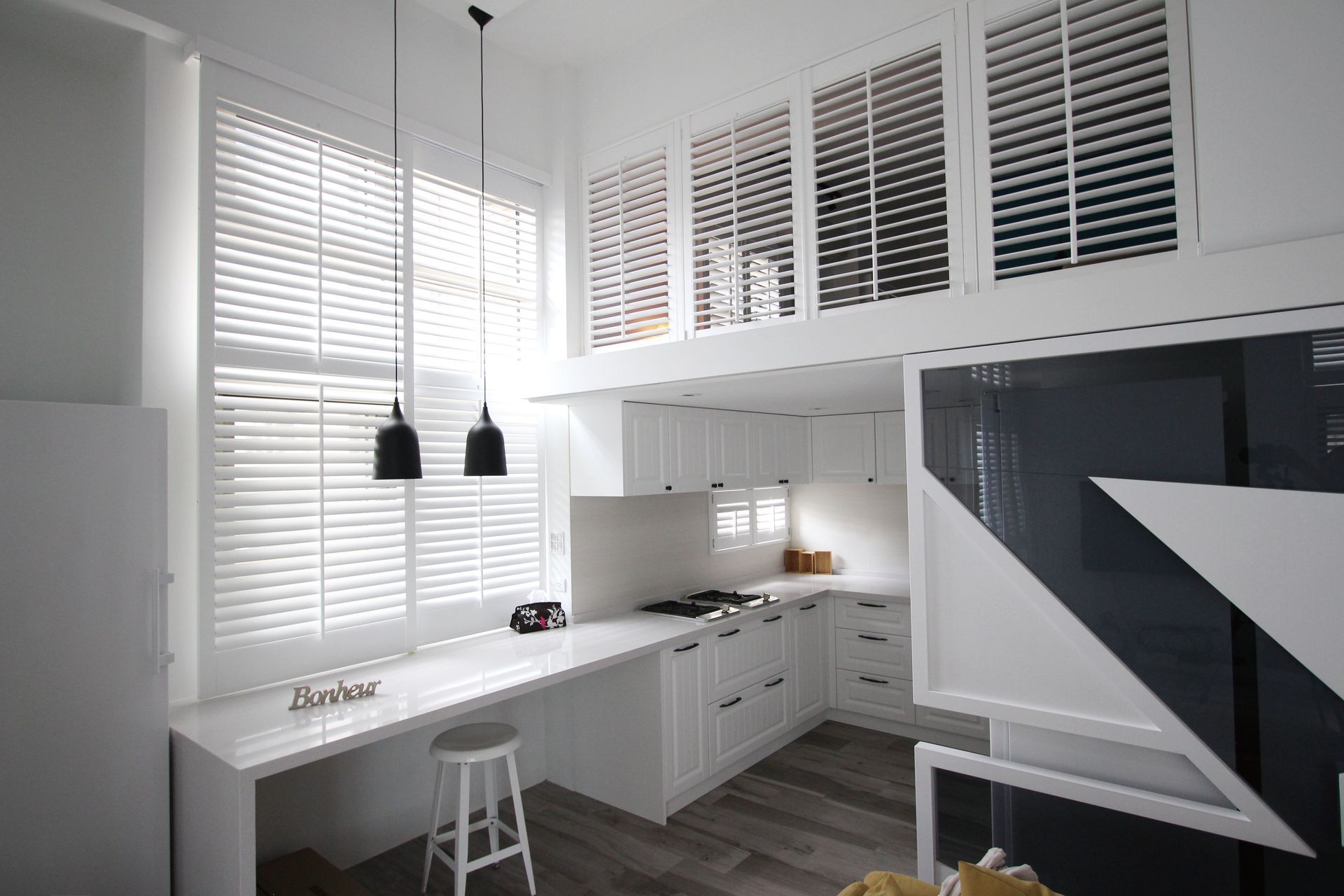 White kitchen with tall windows, built-in cabinets, and a counter with a stool. Black pendant lights hang.