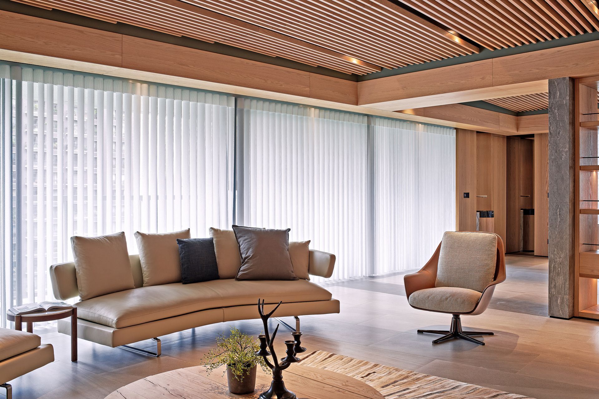 Living room with beige sofa, armchair, and light-colored wood ceiling, and floor-to-ceiling white blinds.