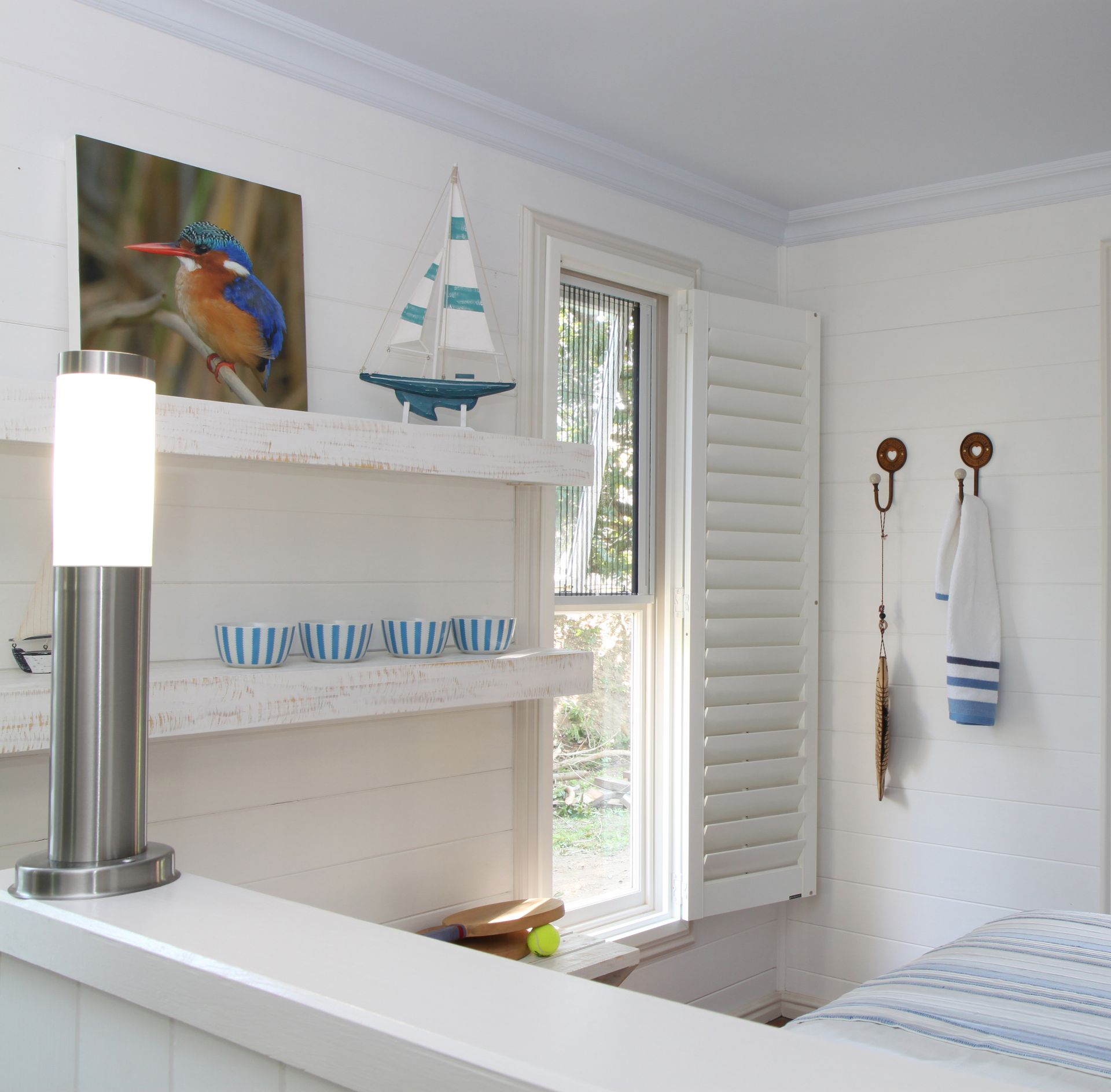 Coastal-themed room with a white shuttered window, shelving, artwork, and a lamp on a counter.