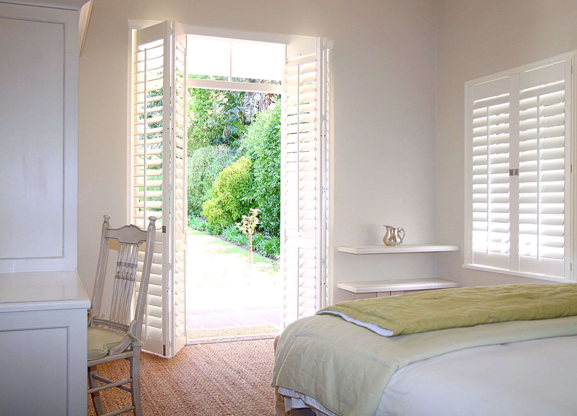 Bedroom with open shutters leading to a garden. White walls, bed, chair, and a rug. Green foliage is visible outside.