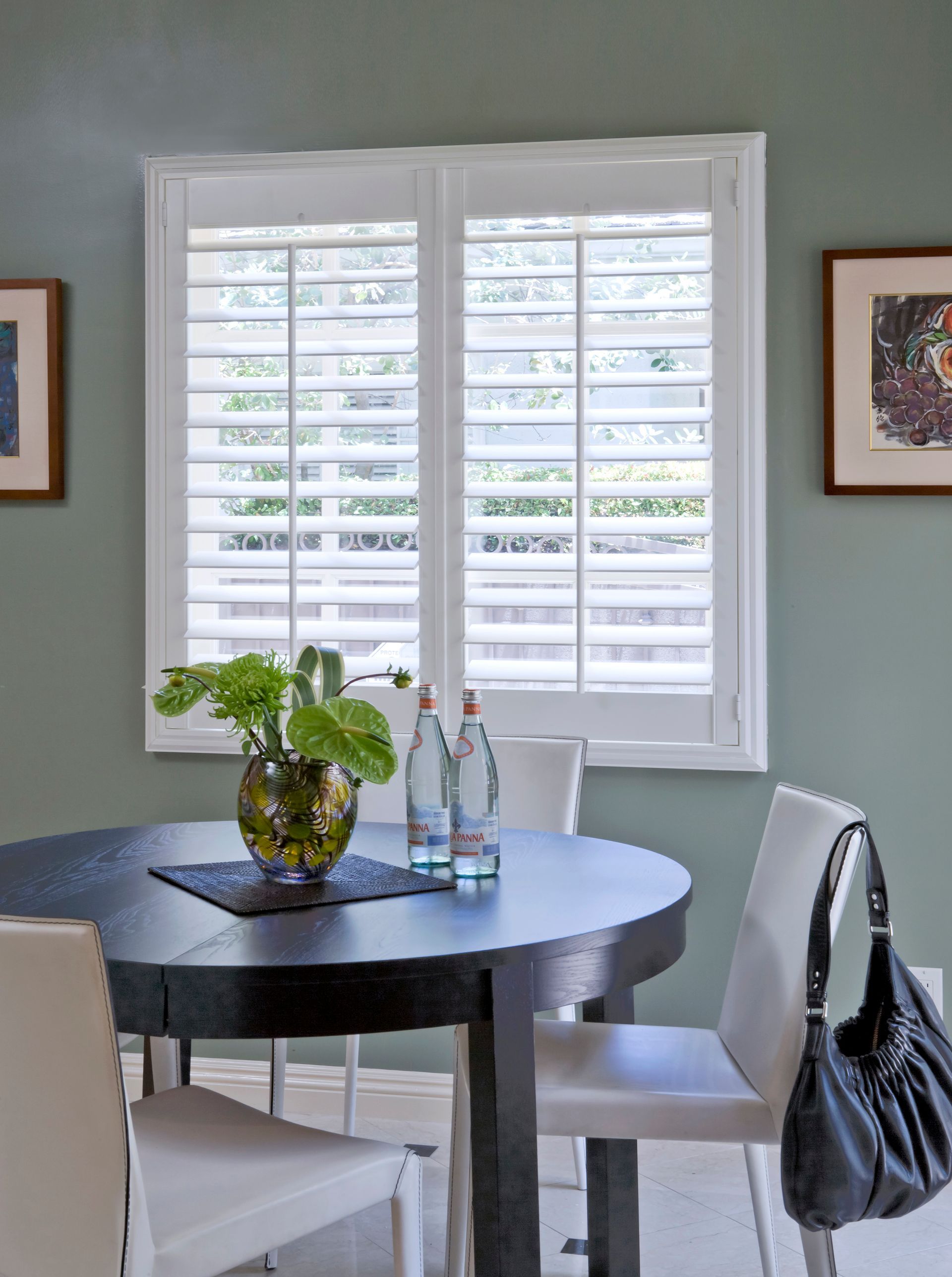Dining room with a round dark table, white chairs, and a window with white shutters.