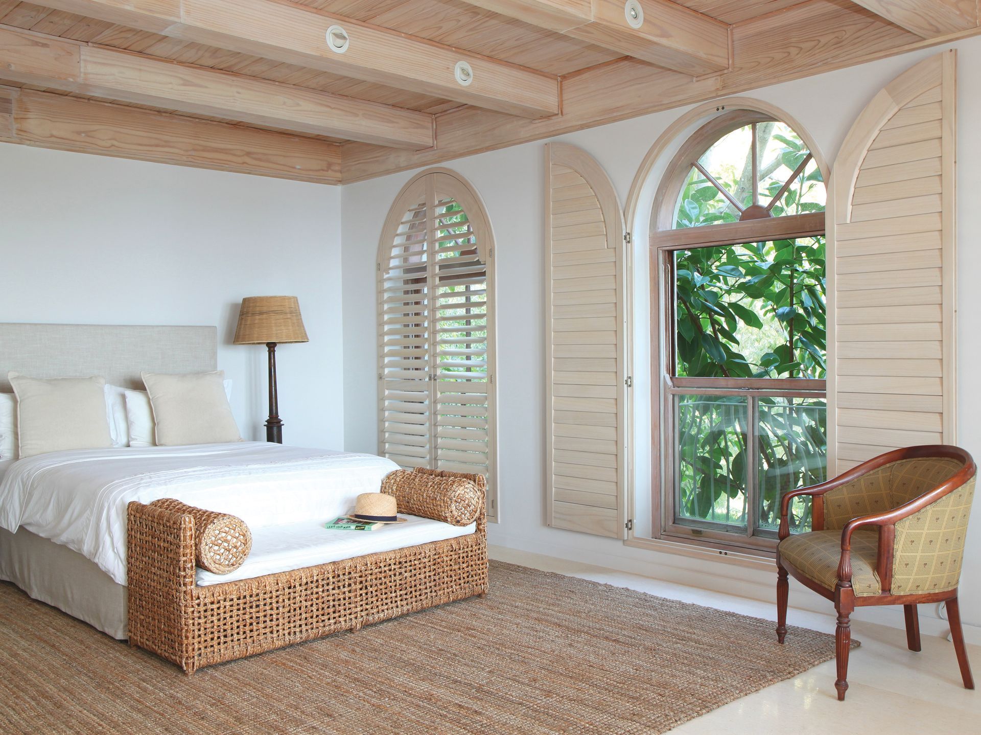 Bedroom with bed, wicker bench, shutters, arched window with greenery, wood ceiling, and chair.